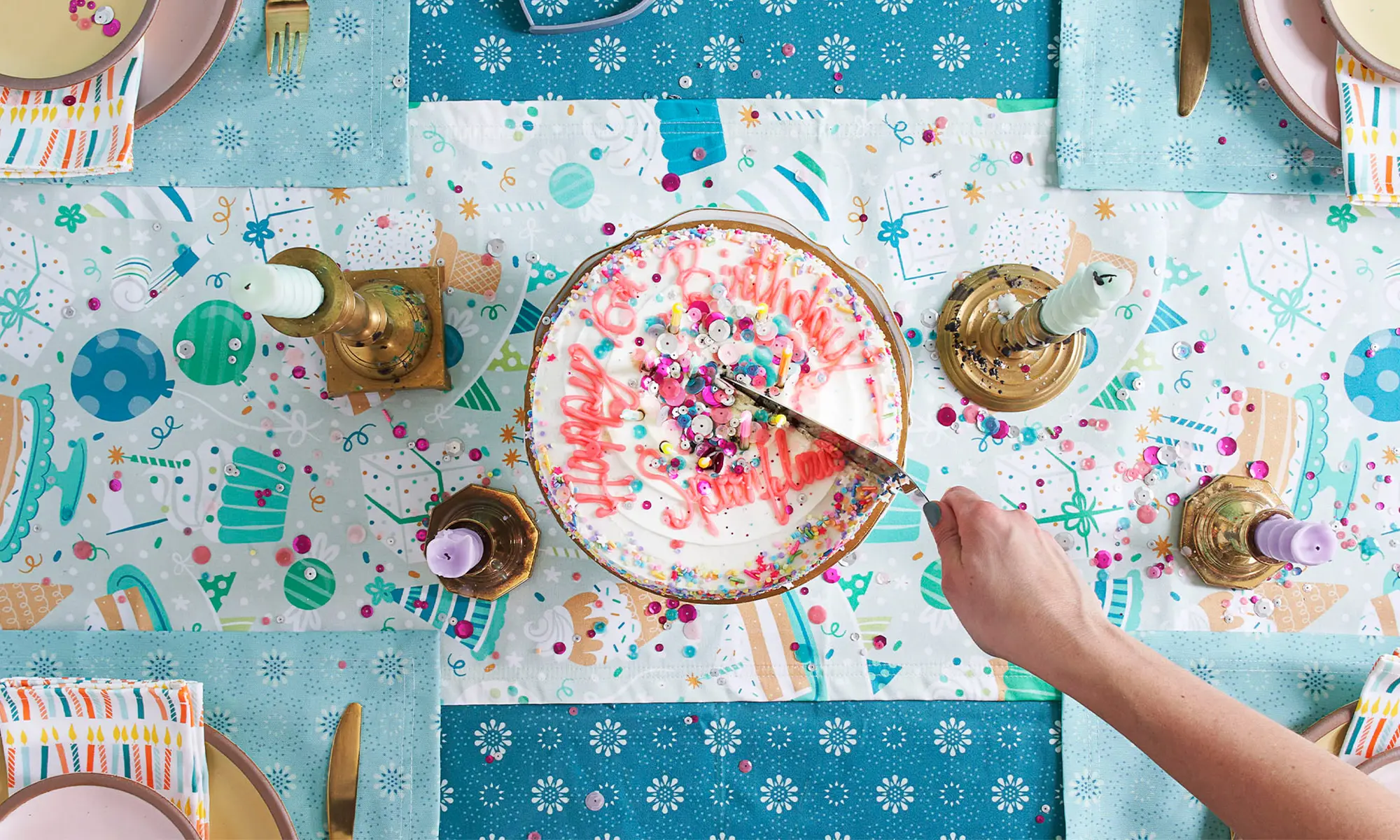 Table with birthday and spoonflower themed runner, tablecloth and placemats and a person cutting a cake.